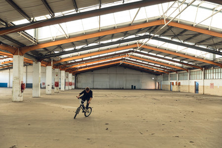 Man performing BMX bicycle stunt inside industrial warehouse wearing dark t-shirt and pants. Stunt, urban, industrial, dynamic, adventure, athletic, machineryの写真素材