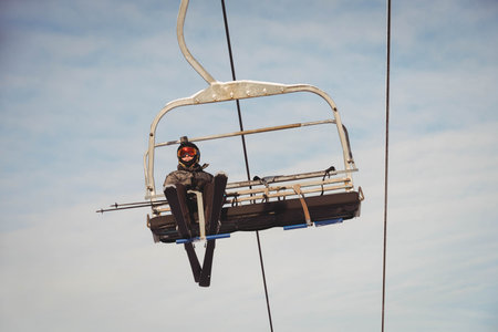 Teenage female skier riding chairlift at ski resort wearing helmet-mounted camera, goggles and skis. Adventure, winter, outdoor, sport, activity, scenic, equipmentの写真素材