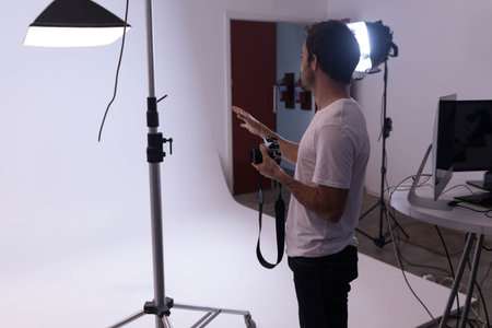 Man holding DSLR camera and gesturing at studio backdrop with softbox light and monitor, copy space. Photography, professional, studio, lighting, equipment, production, creativeの写真素材