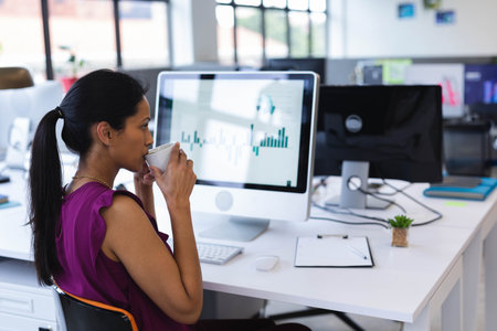 Senior Asian woman analyzing bar charts on monitors and holding coffee mug at office, copy space. Professional, productivity, modern, corporate, workspace, business, technologyの写真素材