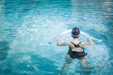 Woman standing chest-deep in outdoor pool wearing swimsuit and cap while holding white foam noodle. Relaxation, aquatic, exercise, outdoorの写真素材