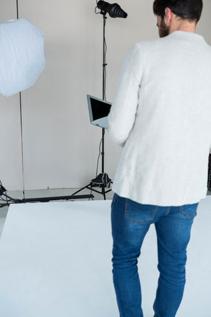 Male studio technician holding laptop before curved backdrop with spotlight and exposed cables. Professional, studio, minimalist, lighting, technology, workspace, commercialの写真素材