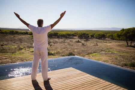 Middle-aged male stretching arms out on wooden pool deck by infinity pool overlooking scrub plain....
