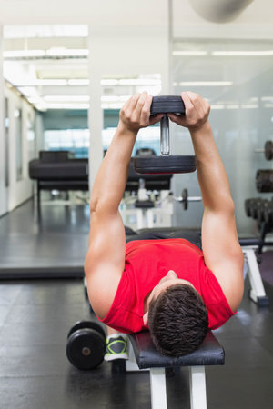 Lying male exerciser in red athletic top on bench in gym holding dumbbell by dumbbell rack. Fitness, strength, workout, training, athletic, health, determinationの写真素材