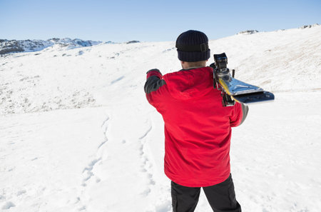 Man wearing red ski jacket, dark beanie carrying skis and poles gazing over snowy mountainside. Adventure, outdoor, winter, activity, athletic, exploration, scenicの写真素材