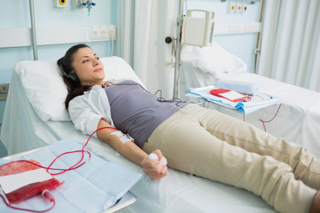 Woman reclining on medical bed at donation center holding stress ball while monitoring IV line. Clinical, wellness, healthcare, relaxation, modern, supportive, therapyの写真素材
