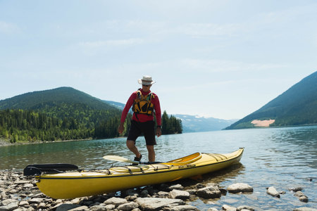 Senior man balancing on yellow kayak at mountain lake shore, holding paddle, wearing life vest, hat. Adventure, outdoor, leisure, nature, tranquility, exploration, scenicの写真素材