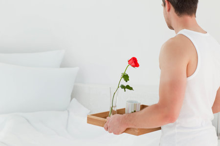 Man standing in bright bedroom holding wooden tray with rose and mug beside bed, copy space. Elegance, serenity, minimalism, wellness, hospitality, sophistication, tranquilityの写真素材