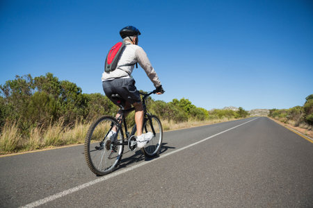 Male cyclist riding mountain bike wearing helmet and hydration pack along rural asphalt road....