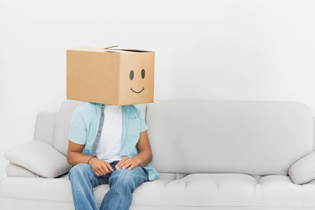 Cardboard box featuring smiley face is sitting on light grey sofa, showing minimal living area. Quirky, humorous, minimalist, relaxation, casual, playful, modernの写真素材