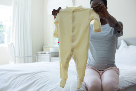 African American woman holding pale yellow footed baby onesie and sitting on bed edge in bedroom. Resilience, motherhood, comfort, tranquility, domestic, cozy, lifestyleの写真素材
