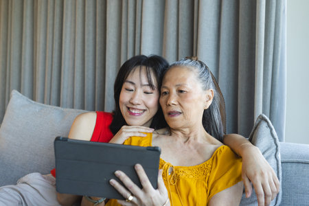 Sitting Asian mother and adult daughter looking at black tablet on grey couch with grey cushion. Familial, generational, bonding, comfort, digital, contemporary, homeの写真素材