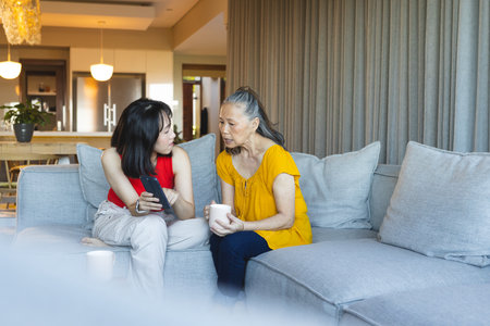 Asian mother and daughter comparing smartphone photos on grey sofa in living room, with ceramic mug. Family, generational, communication, domestic, contemporary, cozy, interactionの写真素材