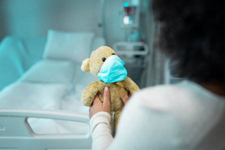 African American woman holding teddy bear wearing blue mask with hospital bed monitor and IV stand. Medical equipment, healthcare, caring, compassion, wellness, supportive, clinical environmentの写真素材
