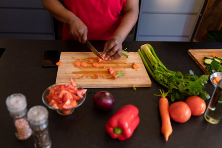 African American mature woman in red shirt slicing carrots on cutting board at kitchen counter....