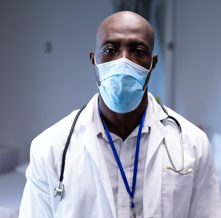 Male doctor wearing surgical mask, stethoscope, lab coat is standing in clinic displaying ID badge....