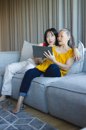Using tablet, Asian mother and daughter sitting on sectional sofa in living room, patterned rug. Family, generational, technology, communication, cozy, contemporary, lifestyleの写真素材