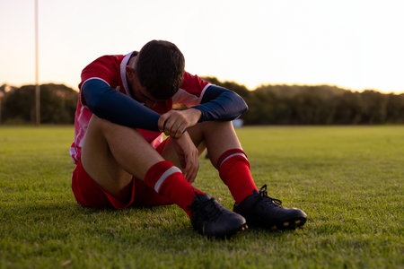 Adult male soccer athlete sitting on grass turf at dusk wearing red jersey and black cleats. Athlete, sports, fitness, outdoor, athleticism, tranquilityの写真素材