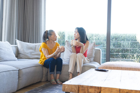 Asian Chinese mother and daughter chatting in bright living room on sofa, holding white mugs. Closeness, warmth, family, companionship, domestic, cozy, lifestyleの写真素材
