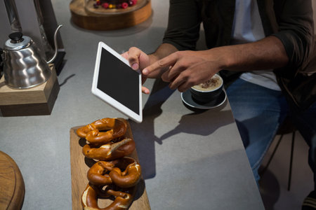 Man wearing jacket tapping tablet at cafe counter with pretzels and cappuccino. Industrial,...