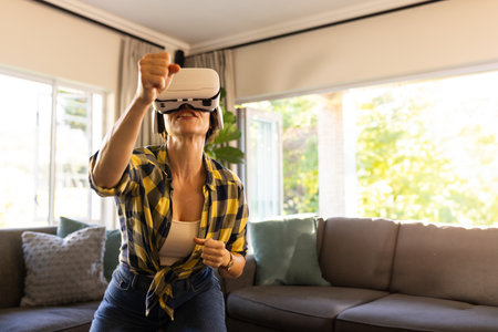 Woman standing in living room wearing VR headset and throwing punch toward grey sofa with pillows. Immersive, interactive, modern, relaxation, leisure, home, vibrantの写真素材