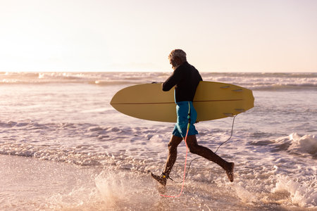 Senior African American man in swim shorts carrying yellow surfboard wading on beach at sunset. Adventure, relaxation, outdoor, scenic, vibrant, active, leisureの写真素材
