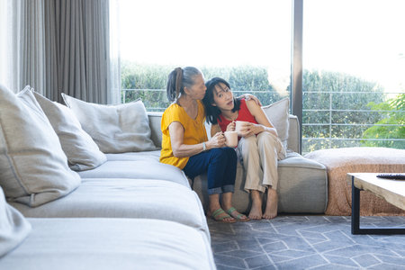 Smiling Asian mother and daughter sitting on sofa in living room, holding white mugs, copy space. Family, companionship, relaxation, cozy, warmth, home, candidの写真素材