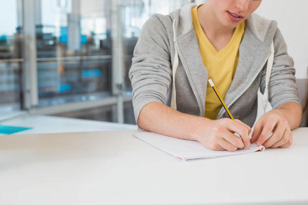 Man wearing grey hoodie writing in notebook with pencil in office with glass partitions, copy space. Student, education, learning, concentration, workspace, modern, professionalの写真素材