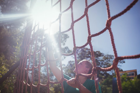 Woman climbing rope net outdoors wearing green athletic shirt and fingerless gloves, copy space. Adventure, fitness, challenge, nature, determination, outdoor, activeの写真素材