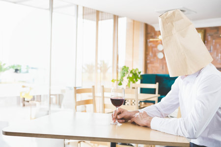 Man sitting at table in cafe holding wine glass with paper bag over head, copy space. Solo, contemplative, rustic, sophisticated, introspection, interiorの写真素材