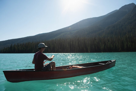 Man paddling red canoe using paddle on turquoise lake while wearing baseball cap T-shirt and shorts. Adventure, serenity, wilderness, exploration, outdoor, landscape, tranquilityの写真素材