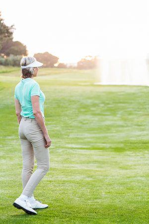 Senior woman standing on golf fairway wearing white visor viewing pond fountain copy space. Leisure, landscape, outdoor, lifestyle, elegance, serenityの写真素材