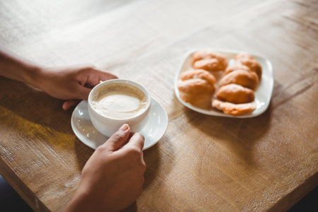 Mid-adult man cradling white ceramic coffee cup and saucer on wooden table with pastries at cafe. Rustic, cozy, breakfast, leisure, minimalism, comfort, morningの写真素材