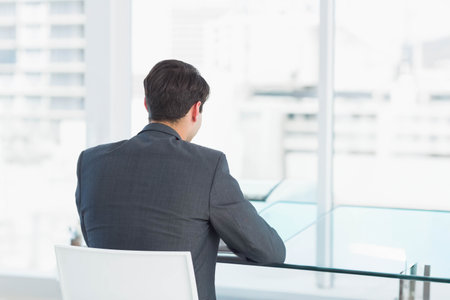 Business professional is sitting at glass-top desk by large windows overlooking skyline in office. Corporate, urban, minimalist, modern, workspace, productivity, executiveの写真素材