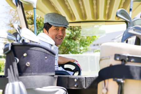 Mid-adult Japanese man sitting in golf cart on fairway wearing flat cap turning head toward clubs. Luxury, leisure, outdoor, landscape, elegance, sport, relaxationの写真素材