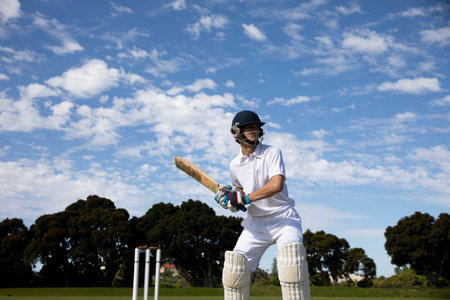 Adult male cricketer wearing helmet standing on green cricket field holding wooden bat, copy space. Athletic, sportswear, dynamic, outdoor, competitive, vigor, preparationの写真素材