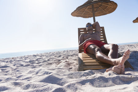 Man reclining on lounge chair under straw umbrella wearing red swim trunks, sunglasses, copy space. Beach, relaxation, leisure, outdoor, vacation, sunny, serenityの写真素材