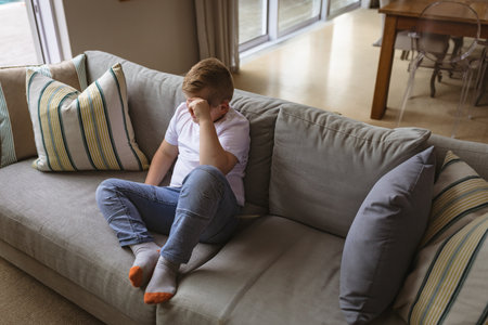 Child boy sitting on light grey sofa with striped cushions at home living room rubbing eyes. Youth, relaxation, cozy, domestic, home interior, leisure, comfortの写真素材