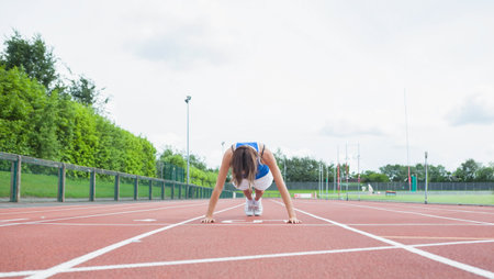 Woman crouching low with hands on white starting line on red track wearing white running shoes. Athletics, sportswear, competition, outdoor, fitness, determination, trainingの写真素材