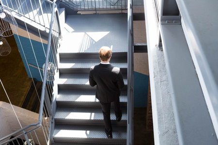Metal staircase rising in industrial stairwell, sunlight casting geometric shadows on steel steps. Industrial, architecture, modern, professional, sleek, urban, geometricの写真素材
