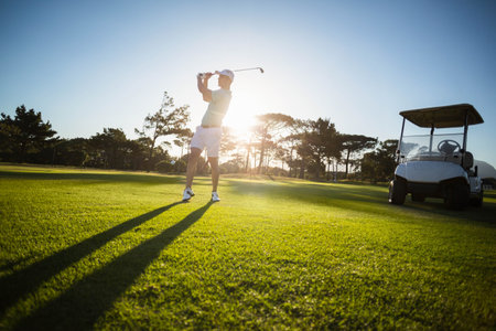 Senior male golfer swinging golf club on fairway during sunset wearing golf attire with golf cart. Athletic, leisure, outdoor, sunset, sport, activity, relaxationの写真素材
