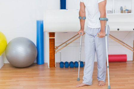 Male patient using forearm crutches standing barefoot in clinic with exercise balls, copy space. Rehabilitation, therapy, physiotherapy, recovery, fitness, wellness, healthの写真素材