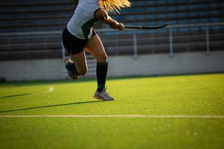 Teenage girl field hockey player sprinting across artificial turf pitch and swinging hockey stick. Athlete, sports, active, training, fitness, outdoors, competitiveの写真素材