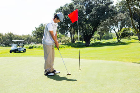 Asian man aligning putter with golf ball on green by red flag with golf cart behind. Athletic,...