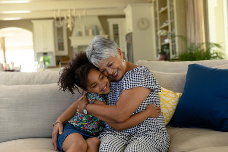 Snuggling senior Hispanic woman and African American girl on sofa in living room with throw pillows. Family, generational, affection, cozy, interior, emotional, bondingの写真素材