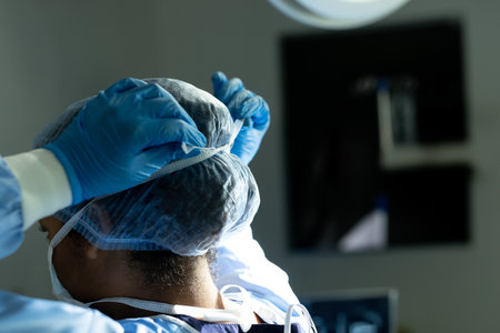 Woman wearing surgical gown pulling cap over head in hospital operating room with light and monitor....