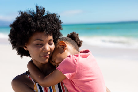 African american woman with eyes closed carrying daughter at beach on sunny day. unaltered, family, lifestyle, togetherness, love and holiday concept.の写真素材