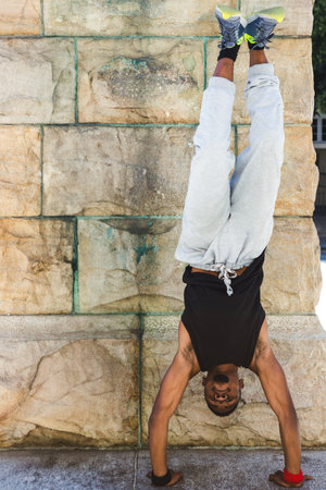 African American man performing handstand against stone wall in plaza wearing running shoes. Flexibility, strength, movement, fitness, outdoor, energetic, dynamicの写真素材