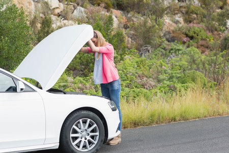 Woman inspecting engine compartment under raised hood of white sedan beside rural paved road. Automotive, maintenance, rural, outdoors, femme, inspection, naturalの写真素材