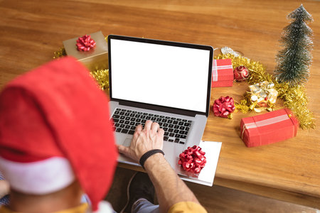 Man wearing red Santa hat typing on laptop at home workspace with holiday gifts and tree. Festive, cozy, workspace, celebration, seasonal, decoration, technologyの写真素材